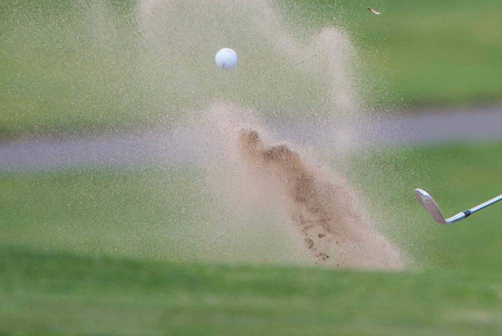 A golf ball soars out of a sand trap as the club head is seen during follow through.