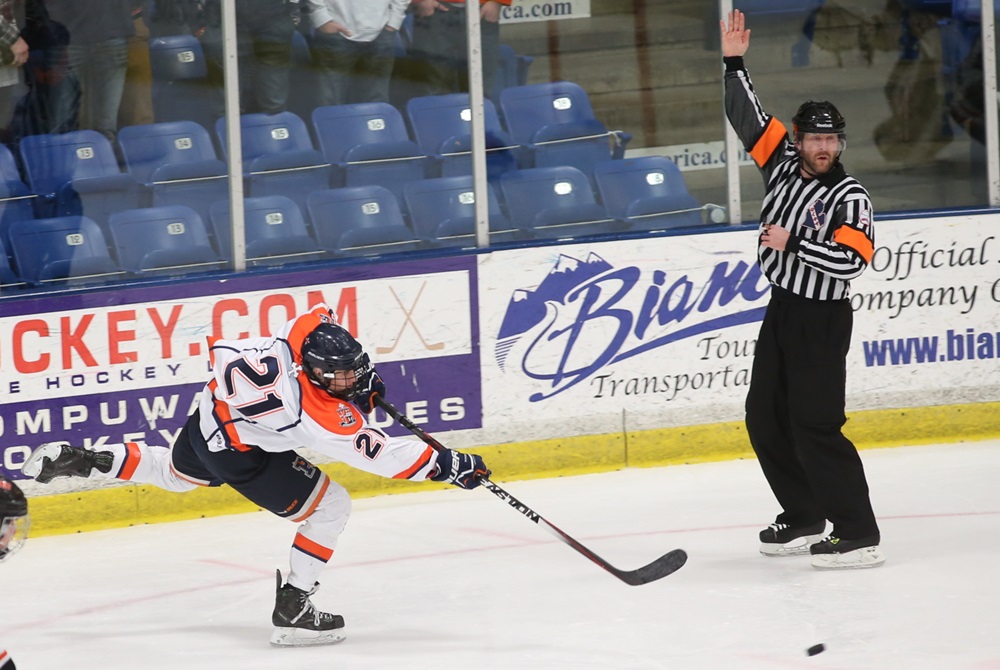 An ice hockey player sends the puck ahead while an official signals a delayed call. 